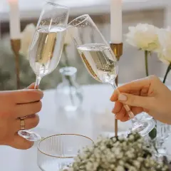 Dwie dłonie wznoszą kieliszki szampana do toastu. Elegancki stół z białymi różami i świecami. Idealny moment, by zastanowić się, ile wina na wesele potrzeba.