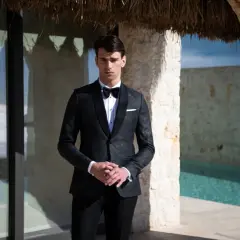 Elegancki mężczyzna w czarnym smokingu, gotowy na black tie event. Stoi przy basenie, otoczony kamiennymi murami.