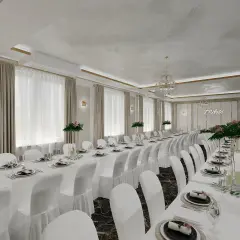 Elegancka, biała sala weselna z długimi stołami nakrytymi białymi obrusami i białymi pokrowcami na krzesłach.