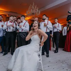 Panna młoda siedzi na krześle, śmiejąc się, otoczona przez weselnych gości i kamerzystę.