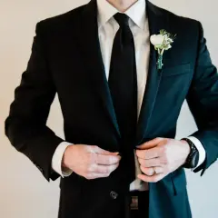 Elegancki czarny garnitur z butonierką, idealny jako jaki garnitur na wesele. Biała koszula i czarny krawat dopełniają stylizację.