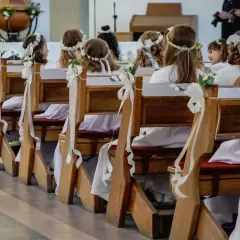Dzieci w bieli, z wiankami na głowach, siedzą w ławkach kościelnych. Czekają na ważny moment, zastanawiając się, ile trwa komunia.