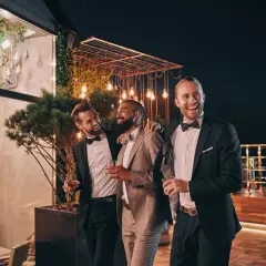 Trzech eleganckich mężczyzn w garniturach, z kieliszkami w dłoniach, śmieje się i rozmawia na tarasie. Jeden z nich, z brodą, jest wyraźnie w roli drużby.