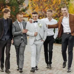 Pięciu panów w eleganckich garniturach, w tym pan młody w jasnym stroju, prezentuje stylowe ślubne cywilne stylizacje na tle jesiennej przyrody.