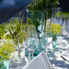 Eleganckie wesele w ogrodzie. Stoły nakryte białymi obrusami, ozdobione zielonymi gałązkami i kieliszkami.