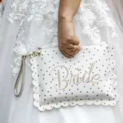 Biała kopertówka w złote kropki z napisem "Bride" to idealny niezbędnik pary młodej. Dopełnia suknię ślubną.