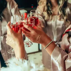 Dłonie w szlafroczkach unoszą kieliszki z czerwonym napojem, na jednej widoczny tatuaż "Bride".