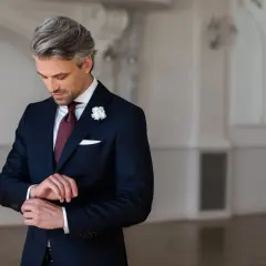 Elegancki strój męski na wesele jako gość: granatowy garnitur, bordowy krawat, biała poszetka i butonierka.
