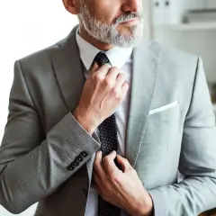 Elegancki mężczyzna poprawia krawat w szarym garniturze szytym na miarę. Cena takiego stroju jest adekwatna do jakości.