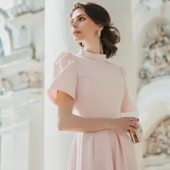 Kobieta w pastelowej sukience z bufkami, trzymająca kopertówkę, na tle eleganckiej architektury.