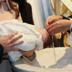 Świadek chrztu podczas mszy trzyma dziecko, podczas gdy kapłan polewa mu głowę wodą.