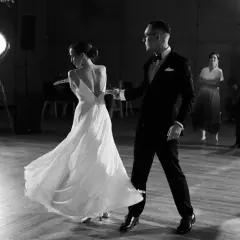Para młoda tańczy, co zamiast pierwszego tańca. Ona w zwiewnej sukni, on w eleganckim garniturze. W tle goście.