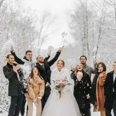 Radosna gromadka rzuca śnieżkami w zimowym lesie. Panna młoda w białym futerku i pan młody w eleganckim płaszczu, otoczeni przez przyjaciół w stylowych okryciach, tworzą piękne stylizacje na wesele zimą.