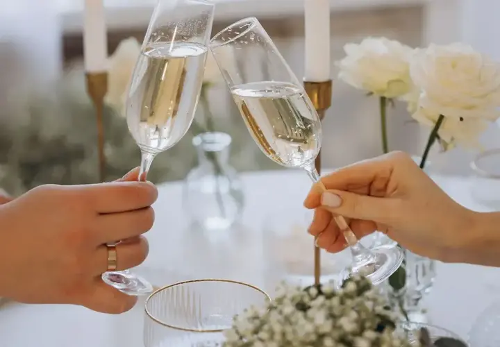 Dwie dłonie wznoszą kieliszki szampana do toastu. Elegancki stół z białymi różami i świecami. Idealny moment, by zastanowić się, ile wina na wesele potrzeba.