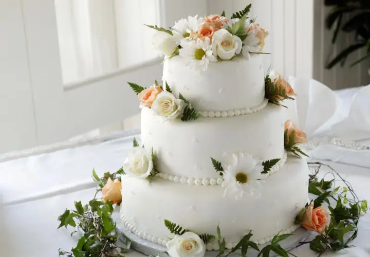 Elegancki tort weselny ozdobiony białymi i brzoskwiniowymi różami, margaretkami i zielonymi liśćmi. Piękne, jakie żywe kwiaty na tort!