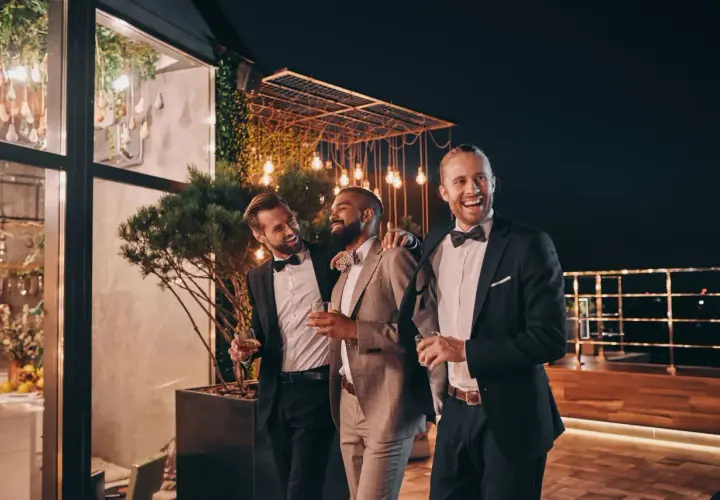 Trzech eleganckich mężczyzn w garniturach, z kieliszkami w dłoniach, śmieje się i rozmawia na tarasie. Jeden z nich, z brodą, jest wyraźnie w roli drużby.