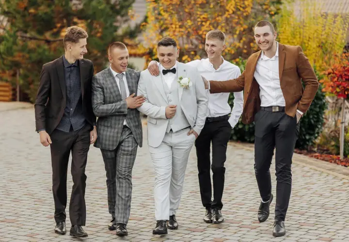 Pięciu panów w eleganckich garniturach, w tym pan młody w jasnym stroju, prezentuje stylowe ślubne cywilne stylizacje na tle jesiennej przyrody.