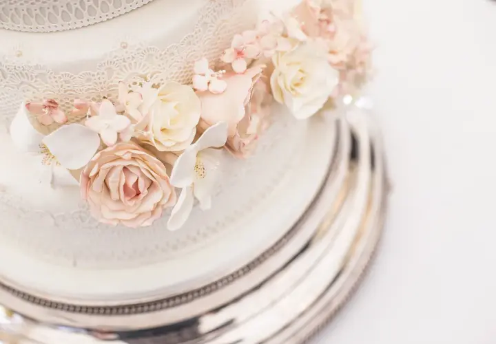 Delikatny tort boho ozdobiony koronką i pastelowymi kwiatami z masy cukrowej, prezentowany na srebrnej paterze.