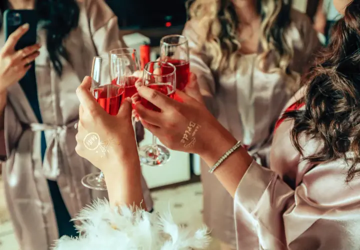 Dłonie w szlafroczkach unoszą kieliszki z czerwonym napojem, na jednej widoczny tatuaż "Bride".