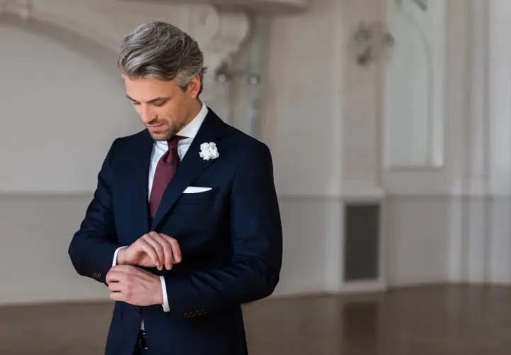 Elegancki strój męski na wesele jako gość: granatowy garnitur, bordowy krawat, biała poszetka i butonierka.