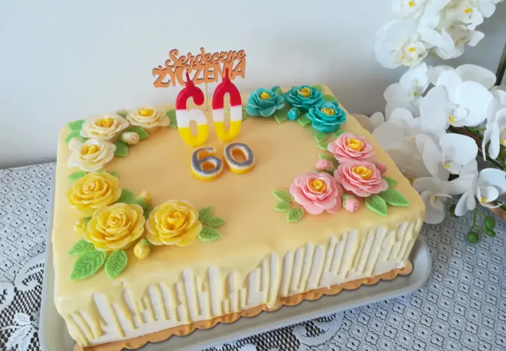Żółty tort z ozdobami kwiatowymi i świeczkami w kształcie cyfr 60.