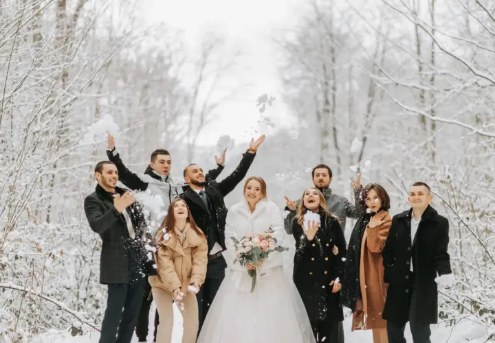 Radosna gromadka rzuca śnieżkami w zimowym lesie. Panna młoda w białym futerku i pan młody w eleganckim płaszczu, otoczeni przez przyjaciół w stylowych okryciach, tworzą piękne stylizacje na wesele zimą.