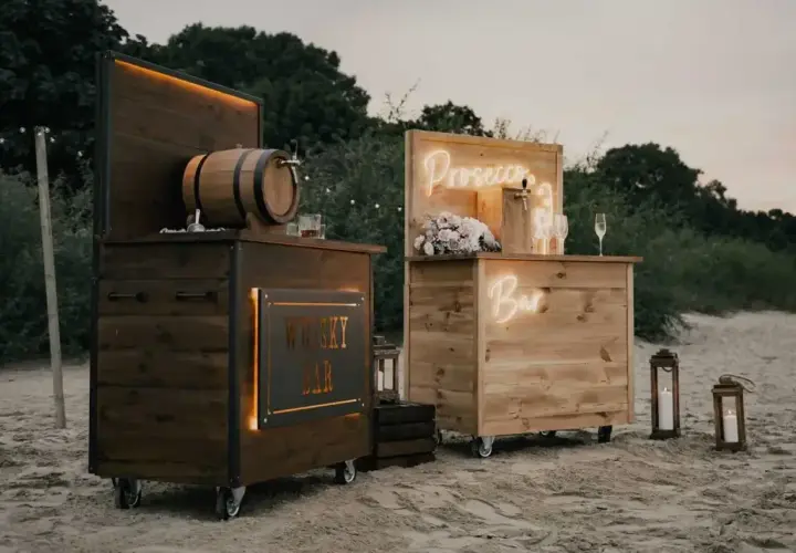 Drewniany shot bar z beczką whisky i neonowym napisem "Prosecco Bar" na plaży.