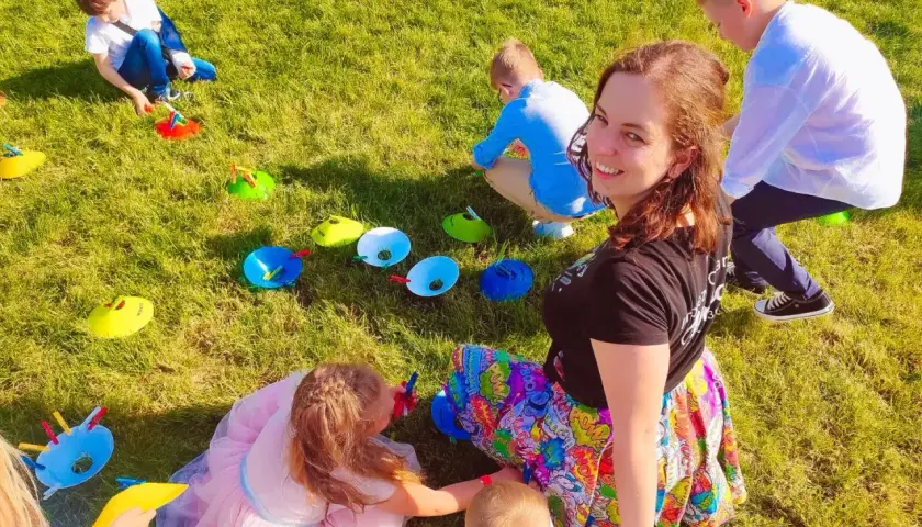 Uśmiechnięta animator dla dzieci cena. Dzieci bawią się na trawie, wykonując zadania z kolorowymi pachołkami.