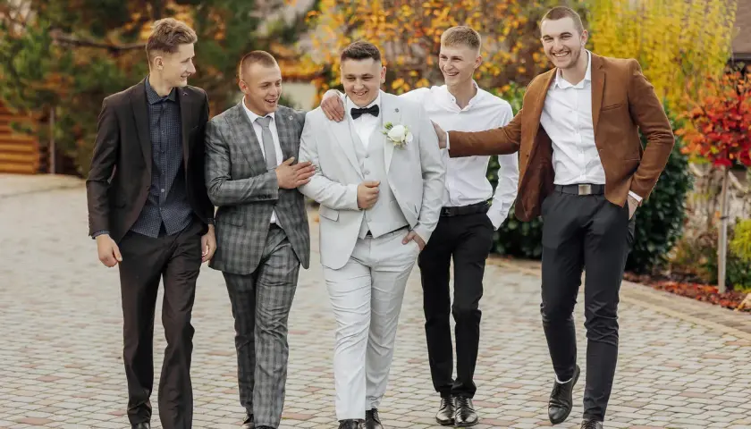 Pięciu panów w eleganckich garniturach, w tym pan młody w jasnym stroju, prezentuje stylowe ślubne cywilne stylizacje na tle jesiennej przyrody.