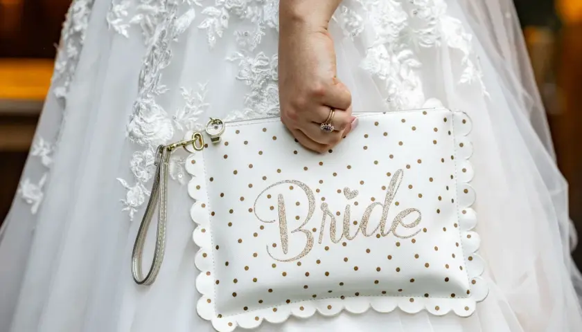 Biała kopertówka w złote kropki z napisem "Bride" to idealny niezbędnik pary młodej. Dopełnia suknię ślubną.