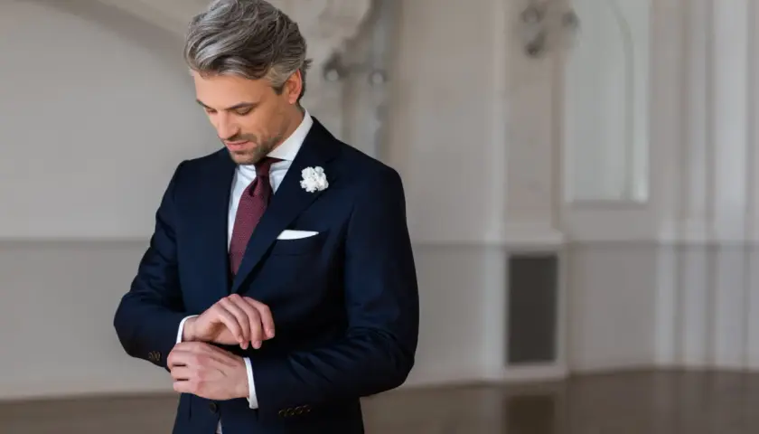 Elegancki strój męski na wesele jako gość: granatowy garnitur, bordowy krawat, biała poszetka i butonierka.