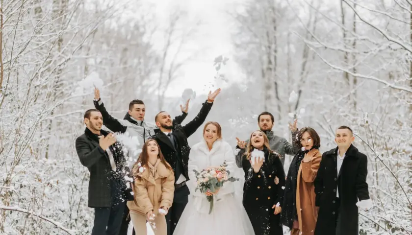 Radosna gromadka rzuca śnieżkami w zimowym lesie. Panna młoda w białym futerku i pan młody w eleganckim płaszczu, otoczeni przez przyjaciół w stylowych okryciach, tworzą piękne stylizacje na wesele zimą.