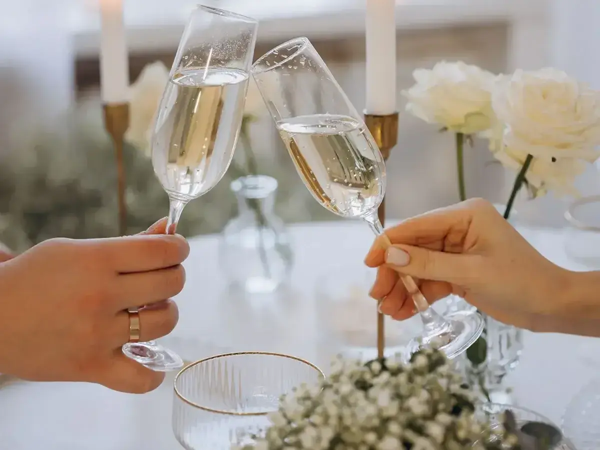 Dwie dłonie wznoszą kieliszki szampana do toastu. Elegancki stół z białymi różami i świecami. Idealny moment, by zastanowić się, ile wina na wesele potrzeba.