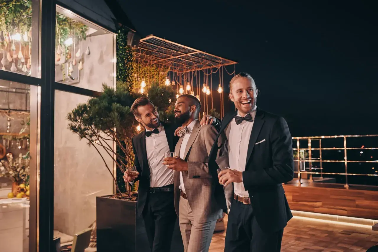 Trzech eleganckich mężczyzn w garniturach, z kieliszkami w dłoniach, śmieje się i rozmawia na tarasie. Jeden z nich, z brodą, jest wyraźnie w roli drużby.