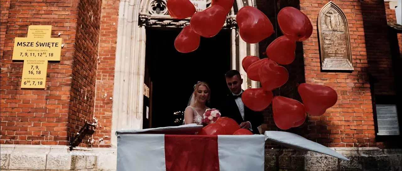 Para młoda wychodzi z kościoła, a rodzice przygotowali niespodziankę: mnóstwo czerwonych balonów w kształcie serc.