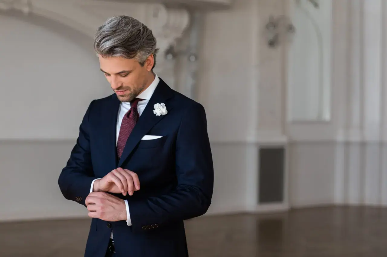 Elegancki strój męski na wesele jako gość: granatowy garnitur, bordowy krawat, biała poszetka i butonierka.