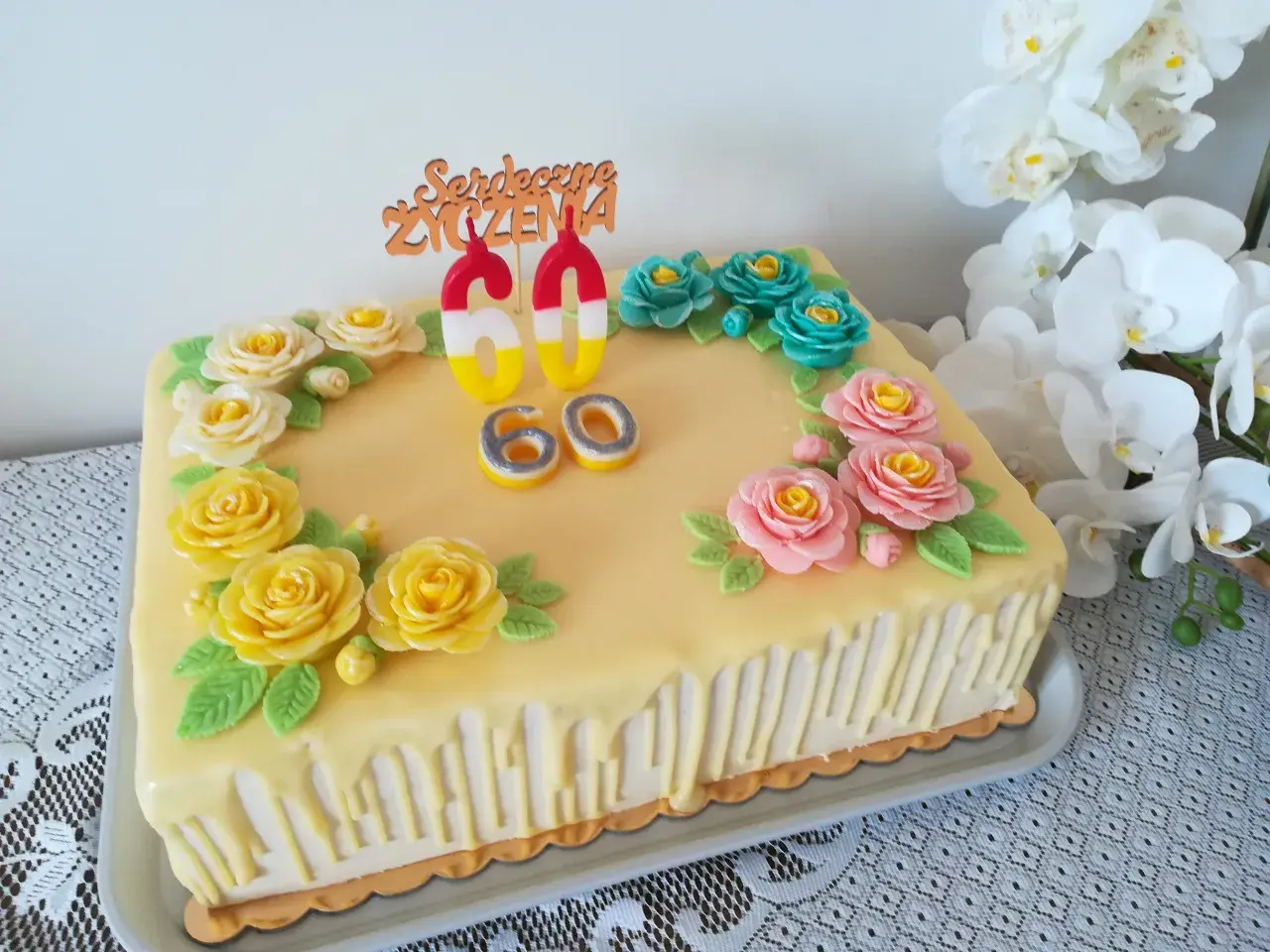 Żółty tort z ozdobami kwiatowymi i świeczkami w kształcie cyfr 60.