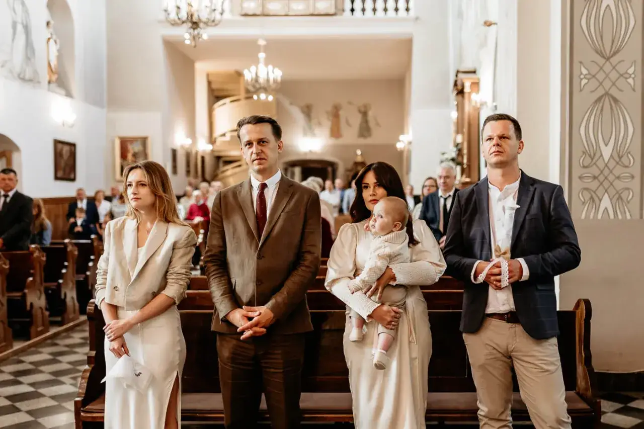 Świadek chrztu podczas mszy, mężczyzna w garniturze, trzyma w dłoniach białą szarfę.