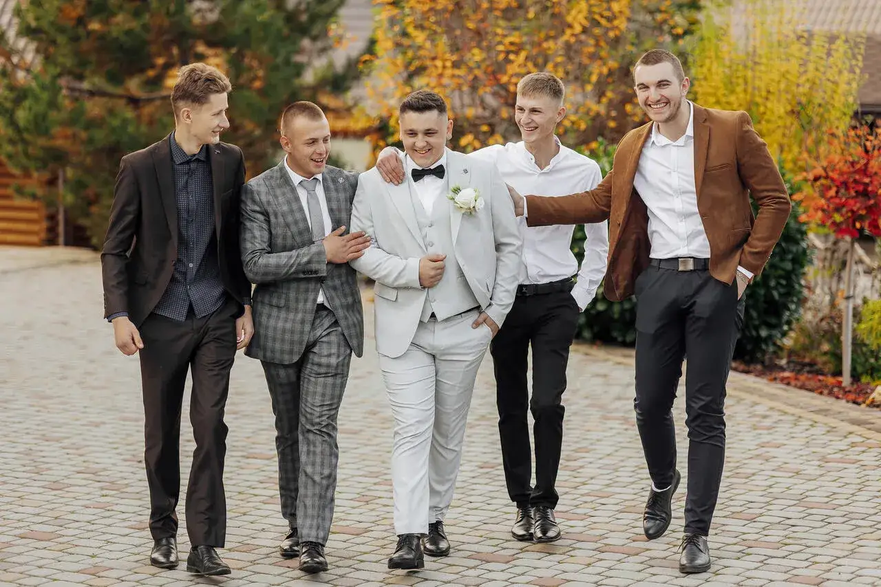 Pięciu mężczyzn w eleganckich strojach, zastanawiających się, jaki garnitur na wesele. Wśród nich pan młody w jasnym garniturze.