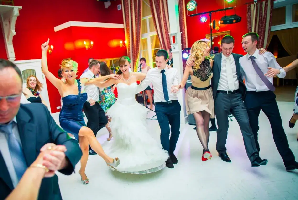 Radosne zabawy weselne na parkiecie. Panna młoda w białej sukni tańczy z gośćmi, wśród nich pan młody i świadkowa w granatowej kreacji.