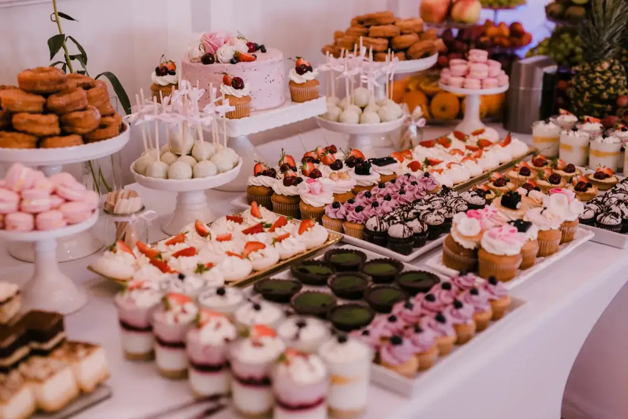 Słodki stół pełen deserów: tort, babeczki, makaroniki, pączki i inne smakołyki, idealne na catering na wesele.