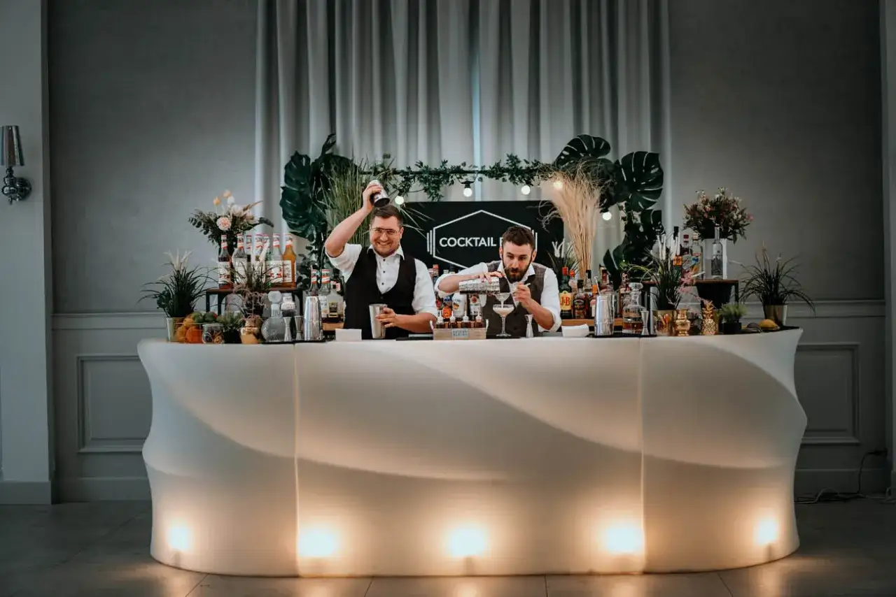 Dwóch barmanów przygotowuje drinki za nowoczesnym, podświetlanym barem. To idealny drink bar na wesele, oferujący orzeźwiające koktajle.