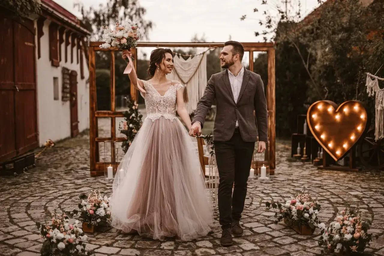 Para weselna idzie przez brukowany dziedziniec, ozdobiony kwiatami i sercem z lampek. Idealne miejsce na fotolustro na wesele.