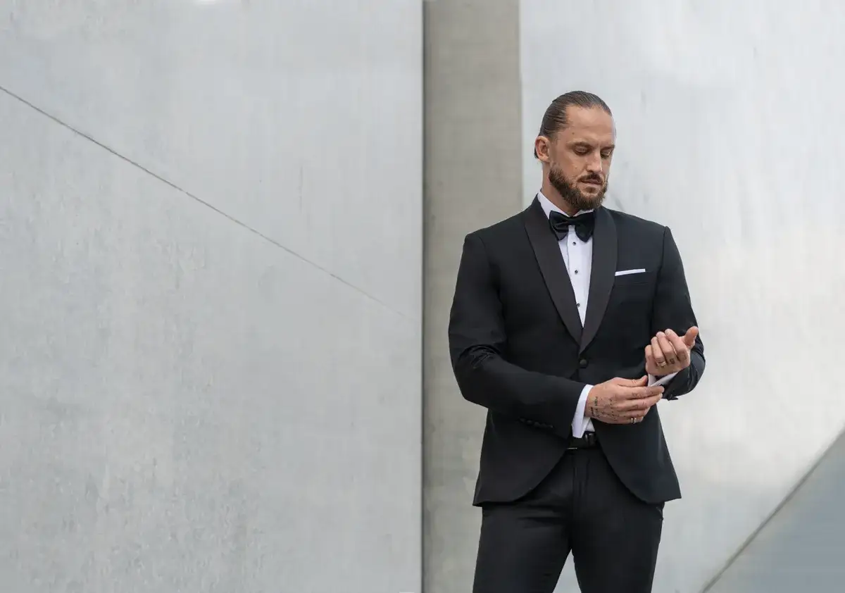 Elegancki mężczyzna w czarnym smokingu z muszką, poprawiający mankiet. Dylemat: krawat czy mucha?