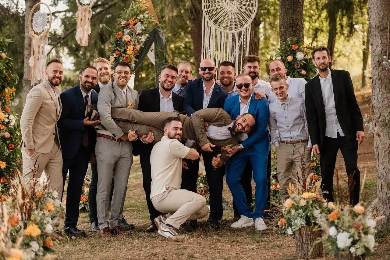 Uśmiechnięta drużba w garniturach, jeden z nich niesiony na rękach, na tle weselnej dekoracji w lesie.