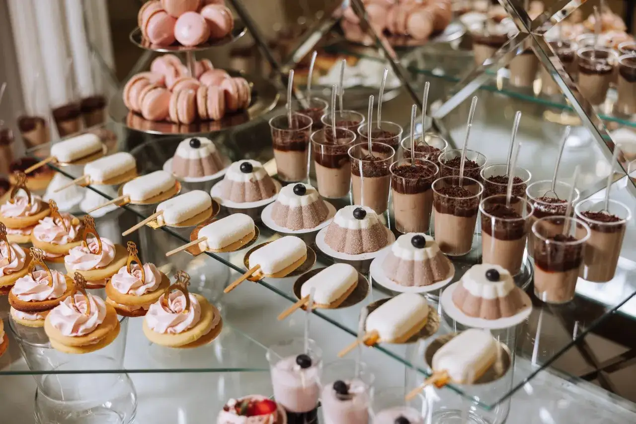 Eleganckie ciasta na wesele: makaroniki, deserki w pucharkach, ciasteczka z różowym kremem i ozdobnymi pierścionkami, lody na patyku.