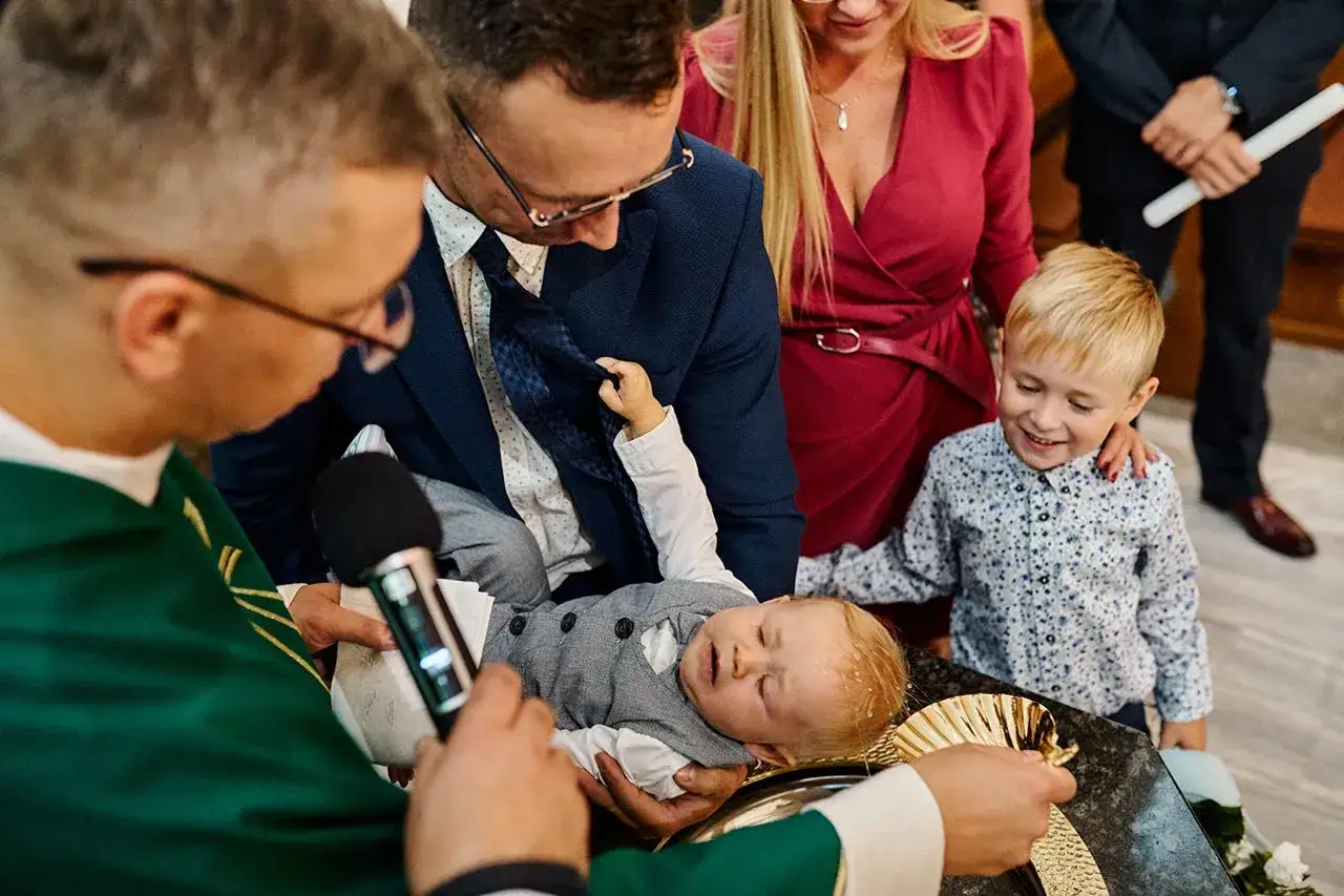 Chrzest dziecka, z dziadkami jako chrzestnymi. Wszyscy patrzą na maluszka podczas ceremonii.