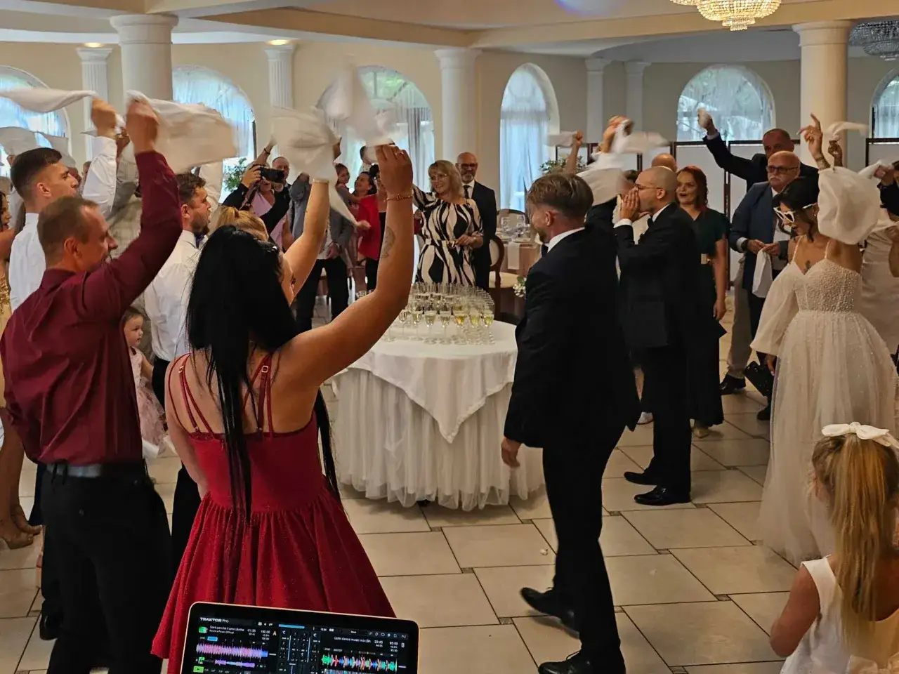 Wesele w pełni! Goście machają chusteczkami, bawiąc się przy muzyce. W tle widać stół z kieliszkami i DJ-a.